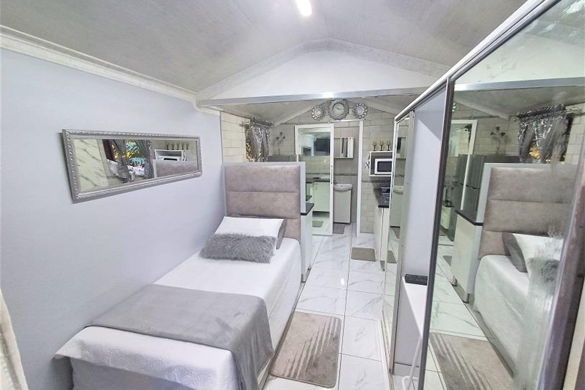A compact bedroom featuring tiled flooring, a vaulted ceiling, and a large mirrored sliding wardrobe, with an adjoining view into a small kitchenette area.