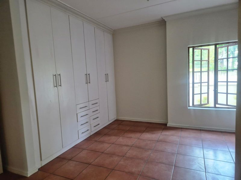A bright, empty bedroom featuring neutral walls, tiled flooring, and extensive built-in cupboard storage with a window providing natural light.