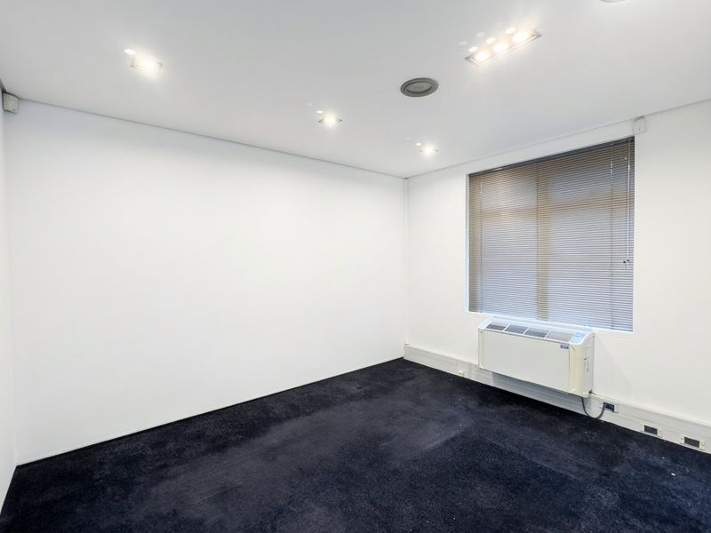 A clean, empty office space featuring recessed lighting, dark carpeting, and a wall-mounted air conditioning unit with window blinds.