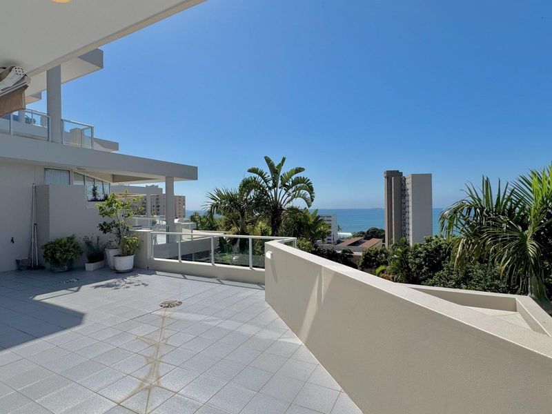 Spacious open-air balcony featuring tiled flooring, a glass balustrade, and a retractable awning, offering expansive coastal and urban views.