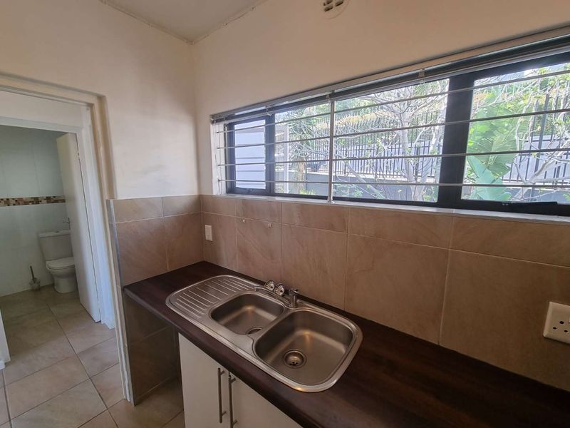 A functional kitchen area featuring a double bowl stainless steel sink, dark laminate countertops, and tiled splashbacks, with an adjacent view into a bathroom.
