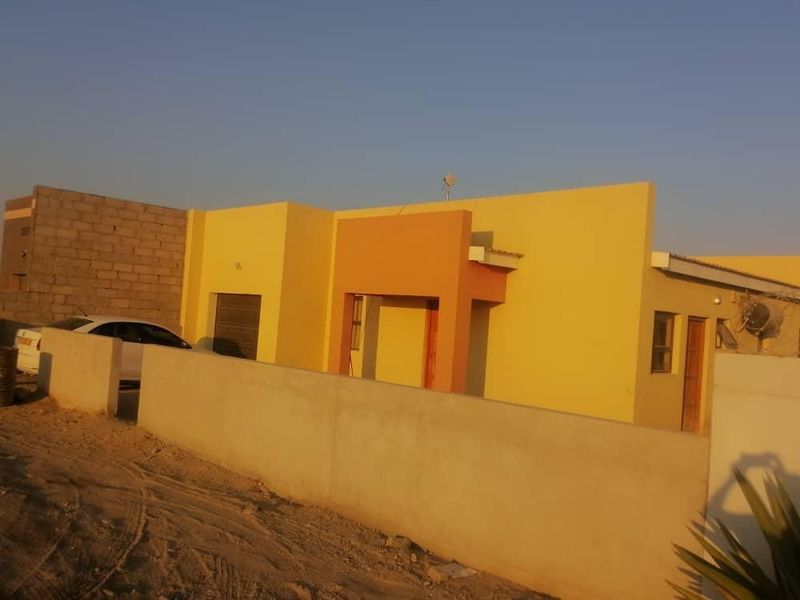 Front elevation of a single-story house featuring a painted facade, integrated garage, and a surrounding boundary wall in a developing residential area.