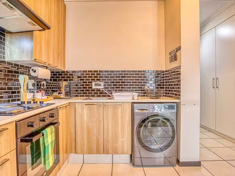 A functional kitchen featuring light wood cabinetry, dark subway tile backsplash, built-in oven, gas hob, and integrated laundry space with tiled flooring.