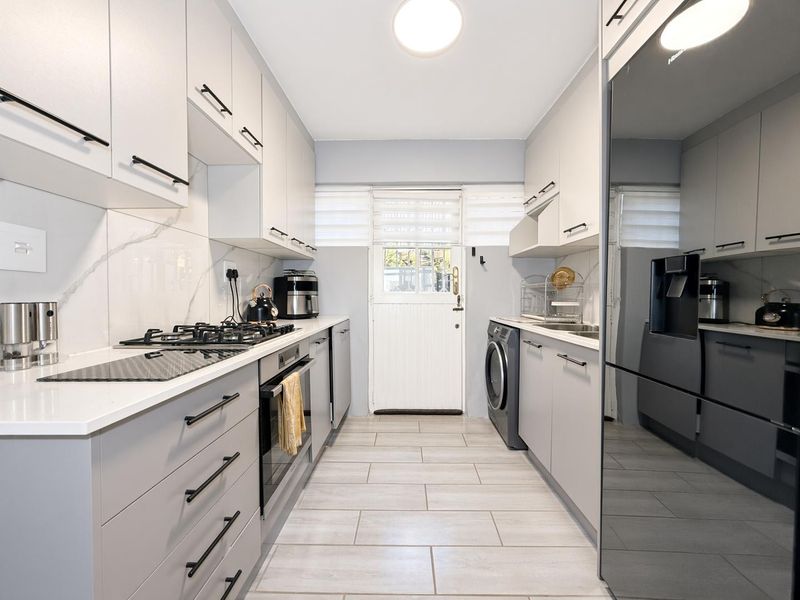 Modern galley-style kitchen featuring sleek grey cabinetry, stone countertops, marble-look splashback, gas hob, built-in oven, and integrated laundry facilities with direct exterior access.