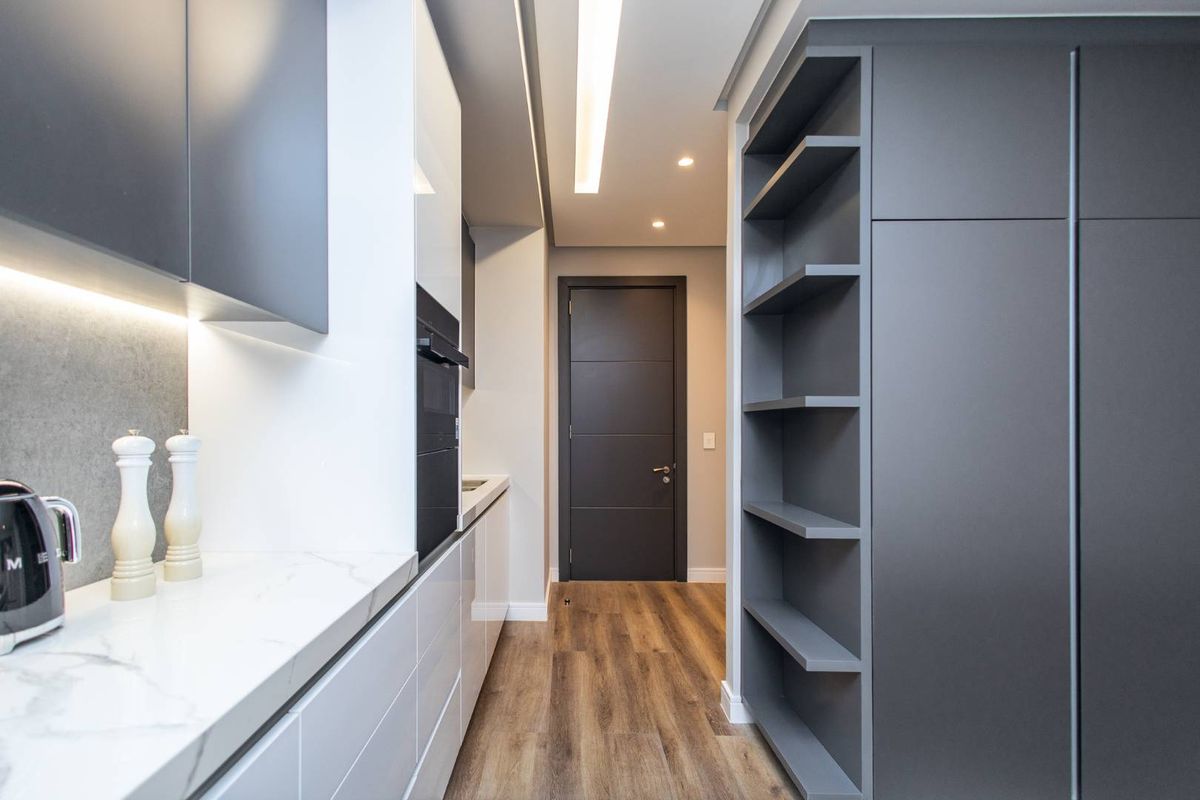 Modern kitchen featuring sleek cabinetry, integrated oven, stone-look countertops, and built-in shelving, leading into a hallway with wood-look flooring.