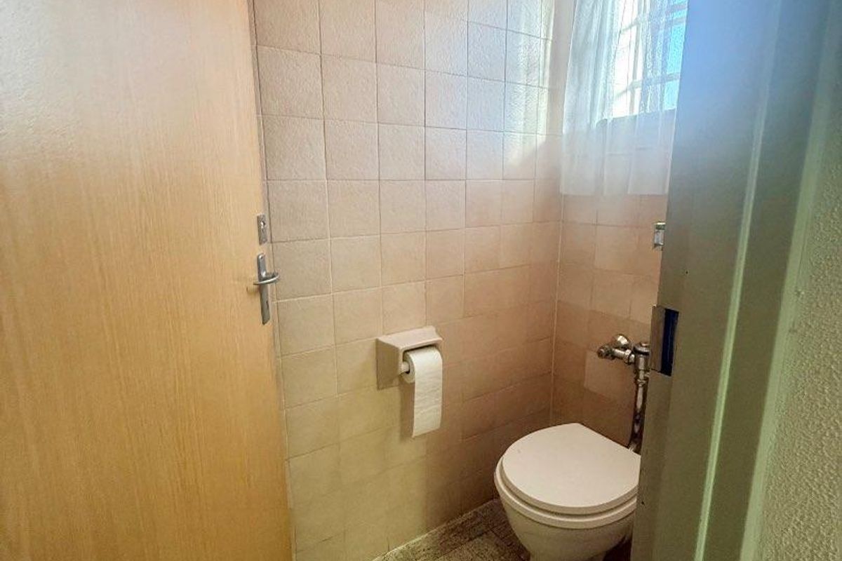 A compact, dated office restroom featuring neutral wall tiles, a standard toilet, and a small window for natural light.