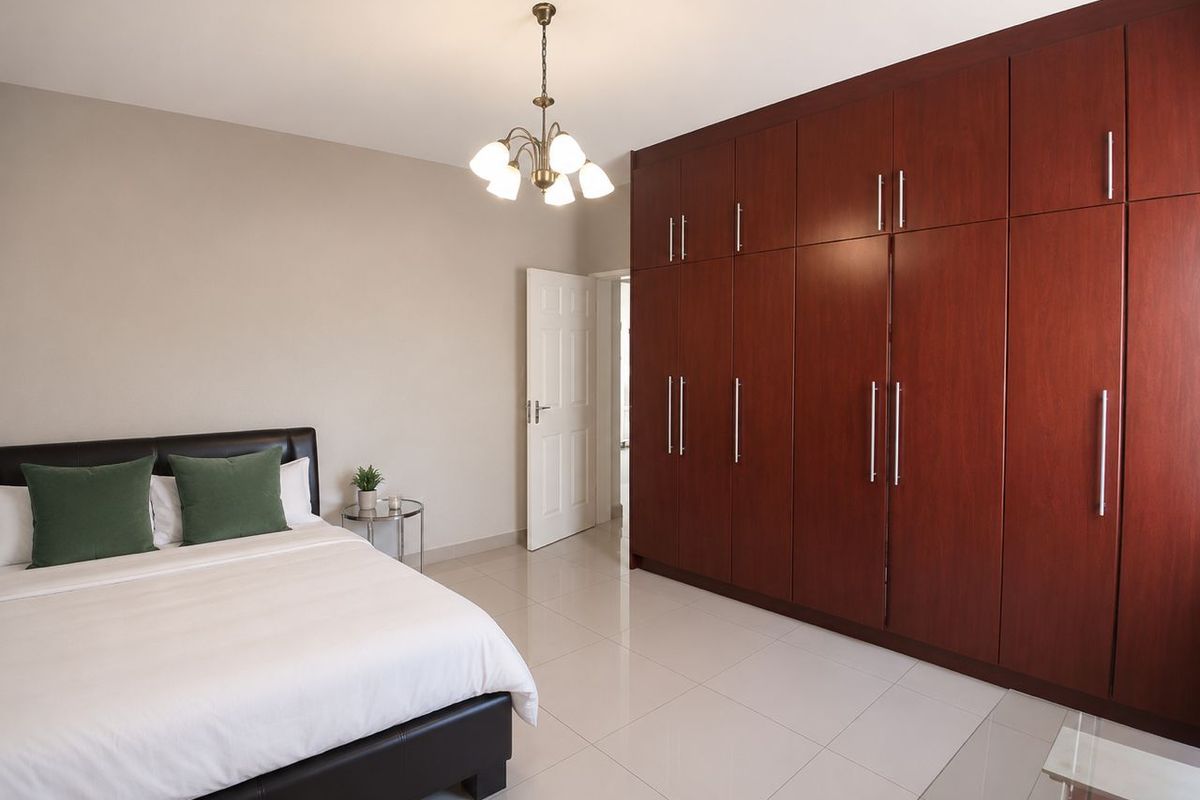 A well-maintained bedroom featuring large, floor-to-ceiling built-in wooden wardrobes, tiled flooring, and a classic chandelier light fixture.
