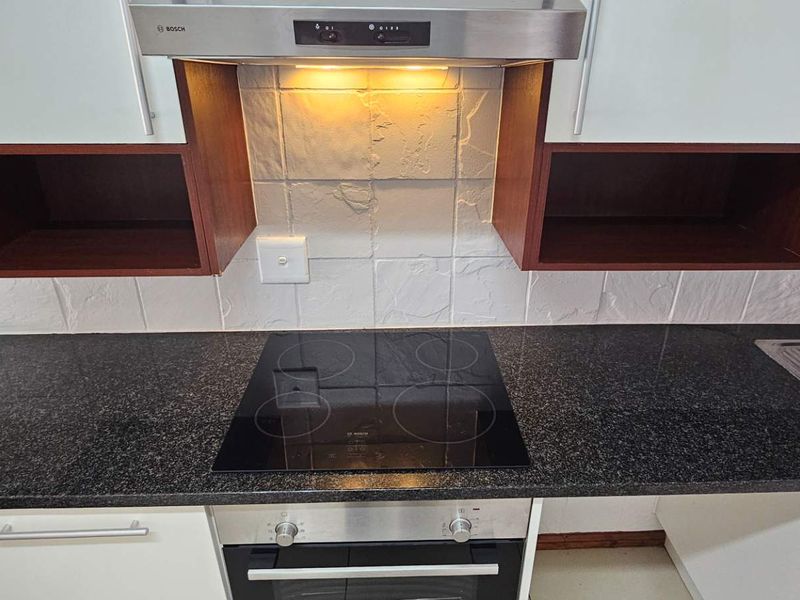 A clean, modern kitchen area featuring a granite countertop, built-in electric hob, oven, stainless steel extractor fan, and integrated sink with textured tile backsplash.