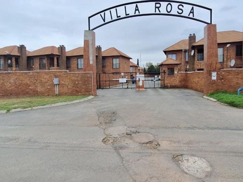 Street view of the Villa Rosa townhouse complex entrance, featuring a brick boundary wall, security gate, and tiled roof architecture.