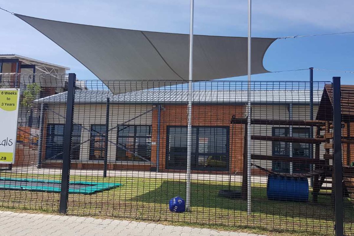 Exterior view of a building facade featuring a shade sail, metal mesh fencing, and a paved sidewalk area.