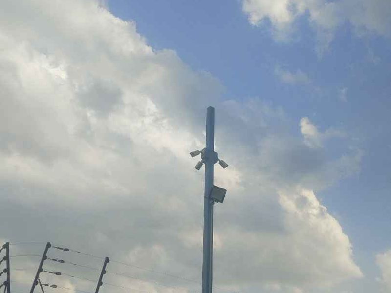 Exterior view showing security infrastructure including electric fencing, a metal pole with mounted security cameras, and a floodlight against a cloudy sky.