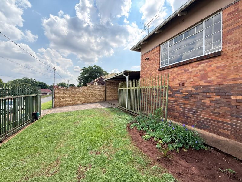 Rear exterior view featuring a brick facade, paved driveway, carport, and secure steel fencing surrounding a small lawn area.
