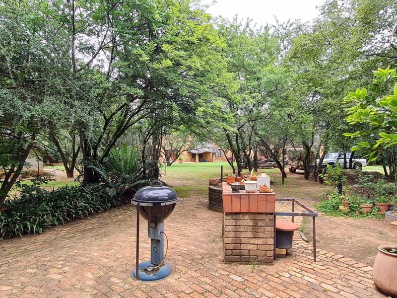 A spacious garden area featuring brick paving and a built-in brick braai, surrounded by mature trees and natural landscaping.