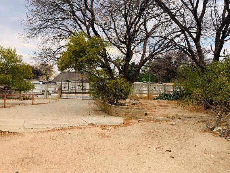 Front exterior view showing a paved driveway, boundary wall with metal gate, and surrounding garden area requiring maintenance.