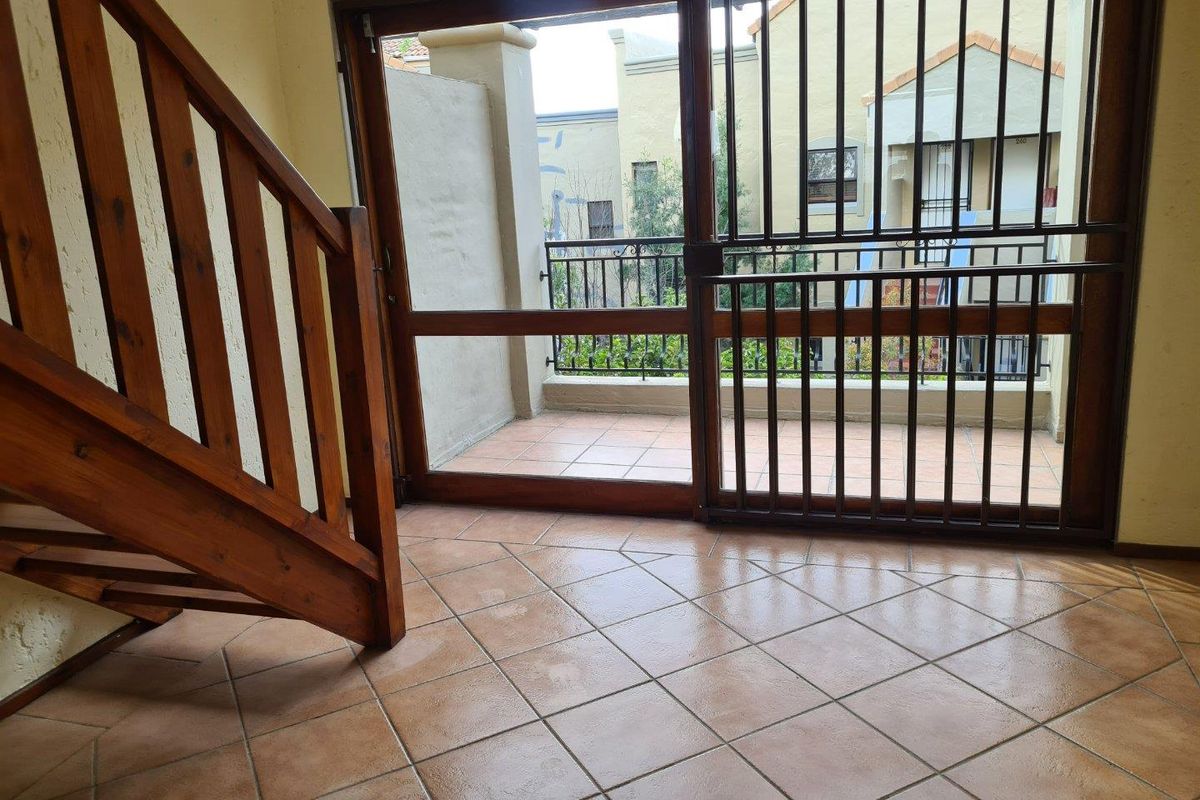 An empty living area featuring tiled flooring, a wooden staircase, and a sliding glass door leading to a private balcony with security gate.