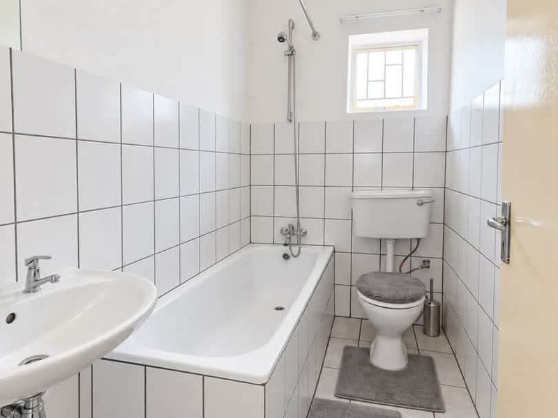 A functional bathroom featuring a white bathtub with shower mixer, pedestal basin, and toilet, finished with classic white wall and floor tiling.