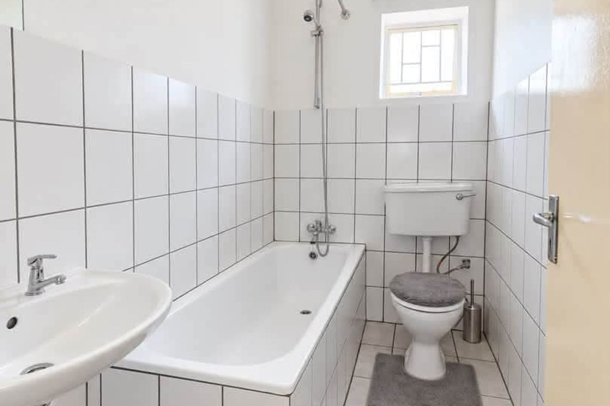 A functional bathroom featuring a white bathtub with shower mixer, pedestal basin, and toilet, finished with classic white wall and floor tiling.