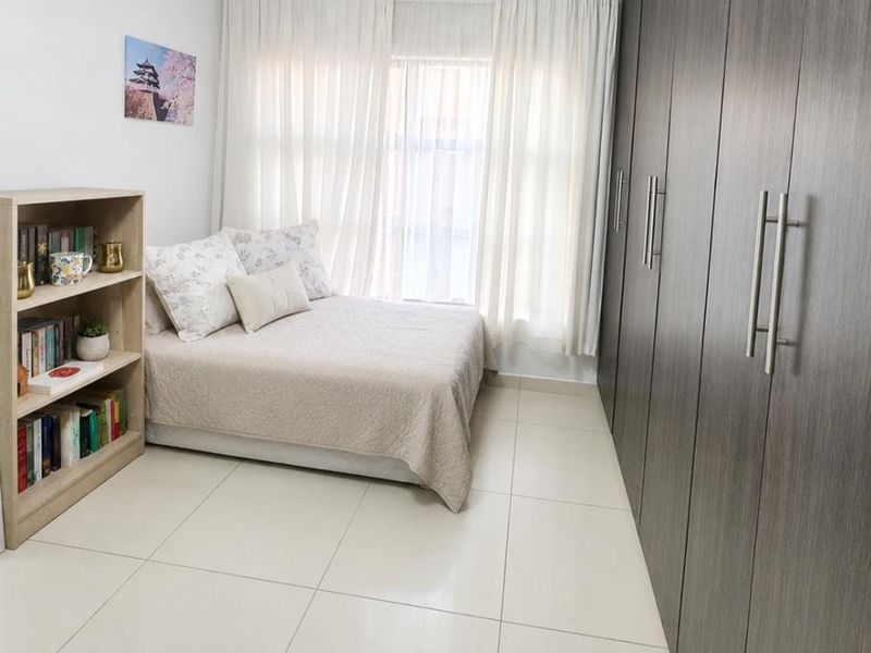A well-maintained bedroom featuring large built-in cupboards, neutral tiled flooring, and ample natural light through window curtains.