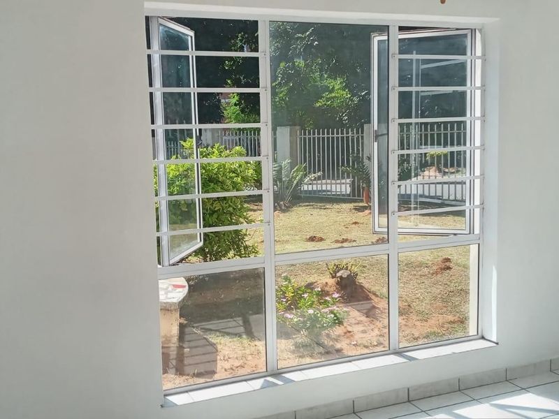 A bright, empty bedroom featuring tiled flooring, a built-in cupboard, and large windows with security bars overlooking the garden.