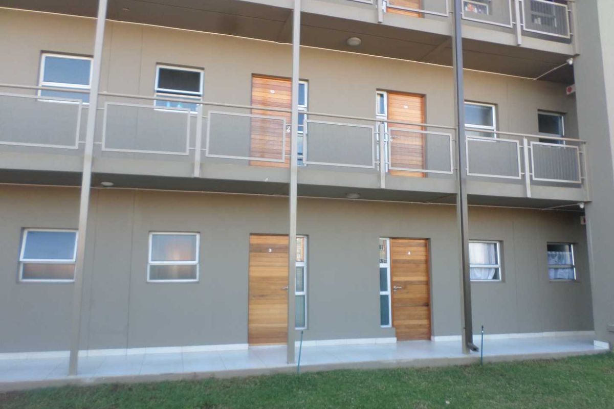 Exterior view of a multi-story apartment building featuring private balconies, timber entrance doors, and a paved walkway overlooking a lawn area.