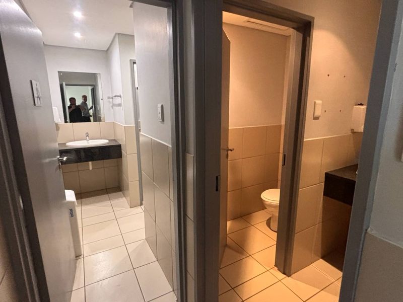 A clean and functional office bathroom facility featuring tiled walls and floors, a vanity basin, and a separate toilet cubicle.