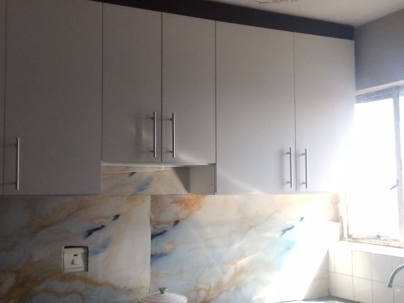 A kitchen area featuring white built-in cabinetry, a marble-effect splashback, and a stainless steel sink with a window providing natural light.