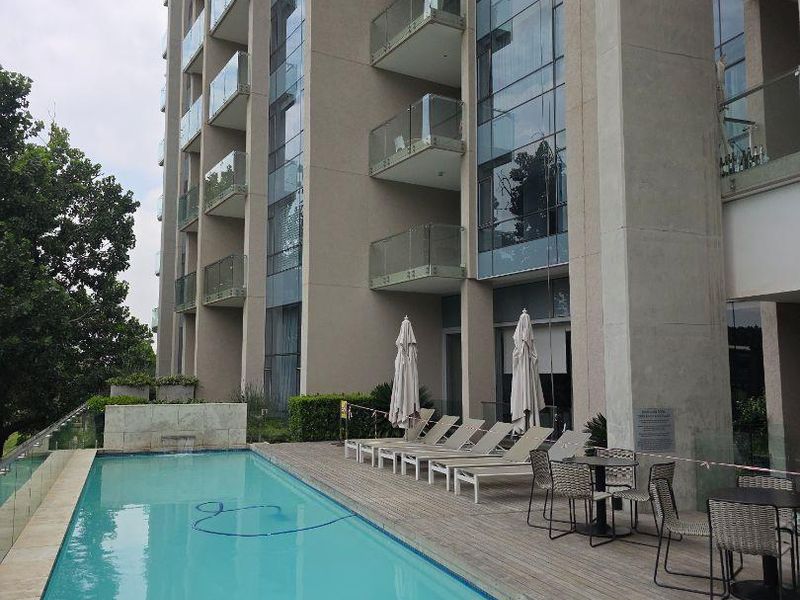 Modern apartment complex pool area featuring a lap pool, timber decking, and glass balustrades with views of the building's glass and concrete facade.