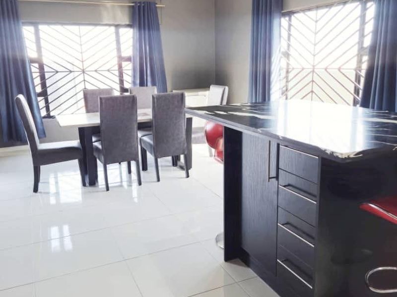 Open plan living and dining area featuring a modern kitchen island with dark cabinetry, polished floor tiles, and large windows with security bars.