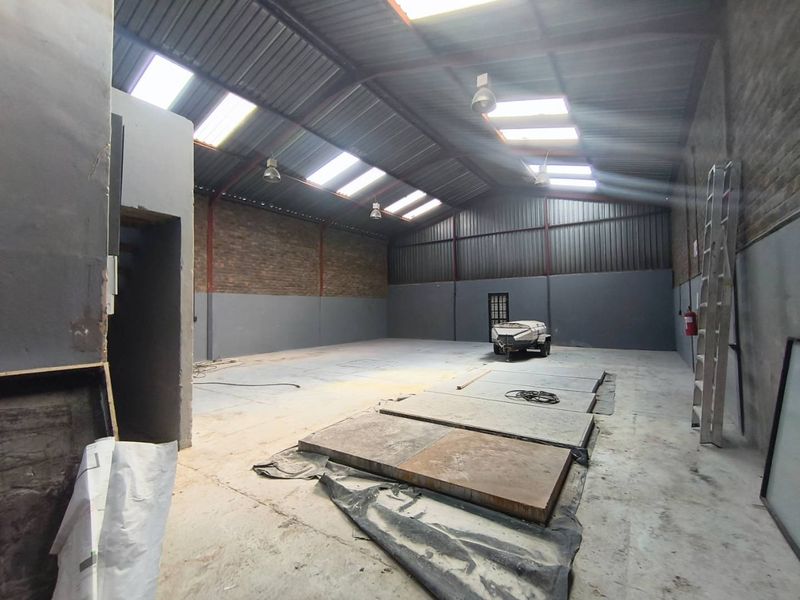 Spacious industrial warehouse unit featuring high ceilings with exposed steel trusses, natural light from skylights, concrete flooring, and exposed brick wall sections.