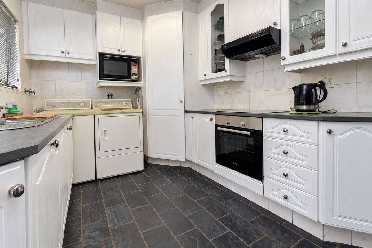 A functional kitchen featuring white cabinetry, dark slate flooring, built-in oven, extractor fan, and integrated laundry appliances with a tiled backsplash.