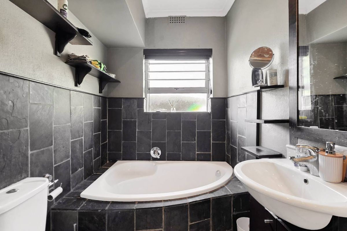 A compact bathroom featuring a corner bathtub with dark slate tiling, a vanity basin, and a louver window for natural ventilation.