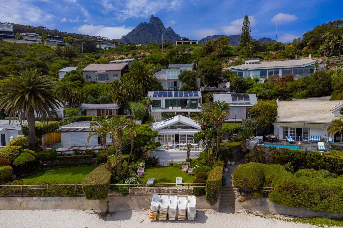 Aerial view of a coastal residential property featuring landscaped gardens, a swimming pool, solar panels, and direct beach access against a mountain backdrop.