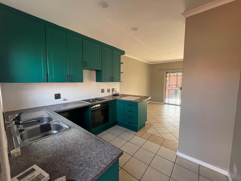 Open-plan kitchen featuring teal cabinetry, laminate countertops, and built-in oven, flowing into a tiled living area with access to a secure exterior door.