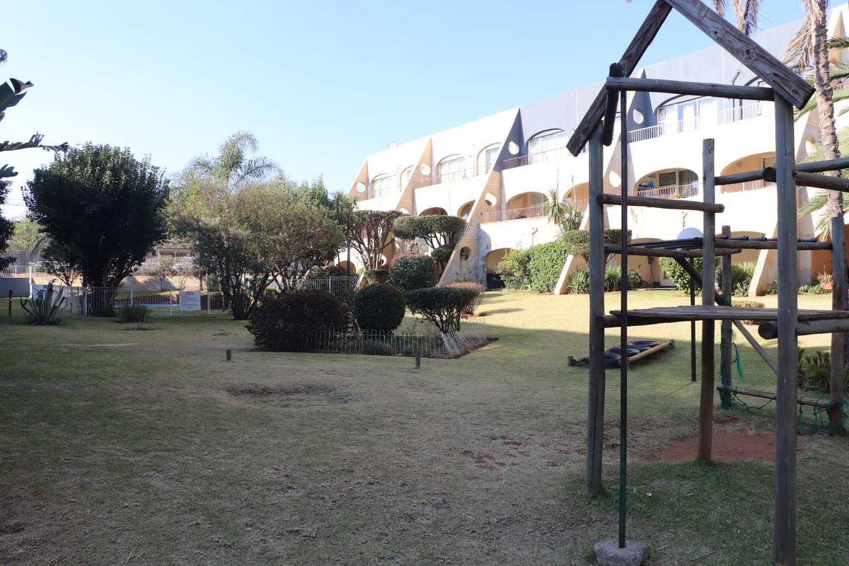A view of the communal garden area featuring a lawn, mature landscaping, and the rear facade of the apartment complex with balconies.