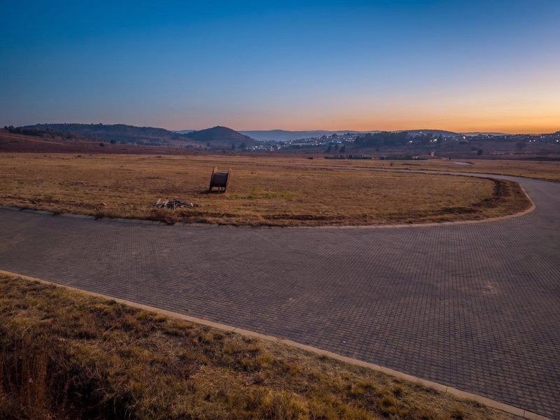 An aerial view of a vacant residential plot featuring paved road access and scenic landscape views at sunset.