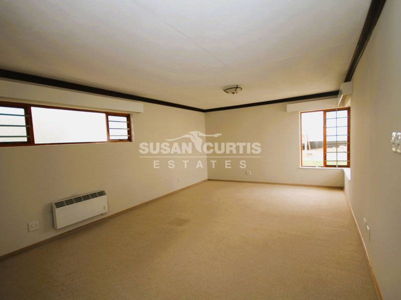 A spacious, empty living room featuring neutral walls, carpeted flooring, timber-framed windows, and a wall-mounted electric heater.