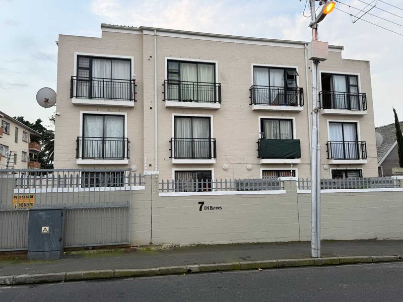 Front elevation of a multi-story apartment building featuring balconies with metal railings, sliding glass doors, and a secure perimeter wall.