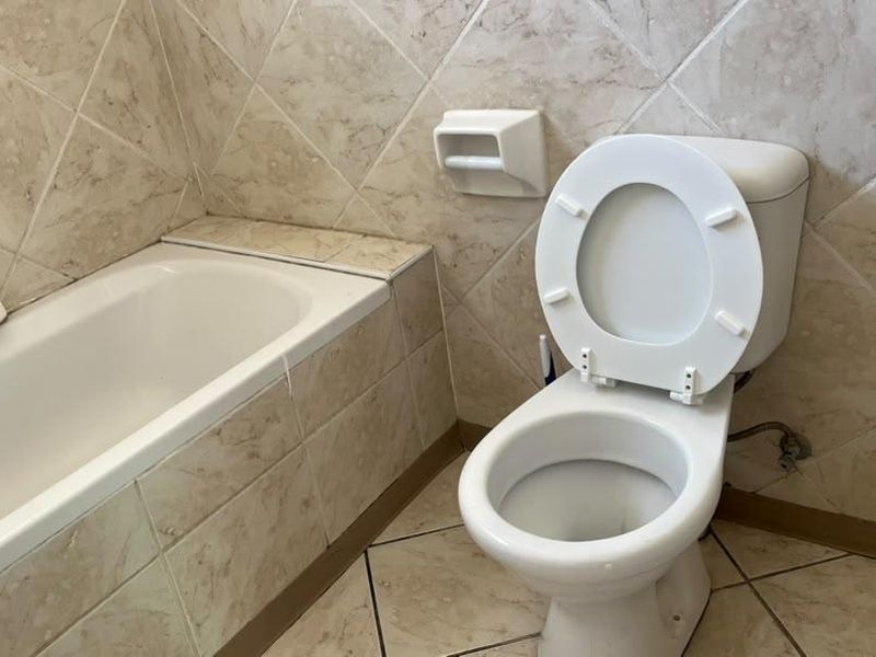 A functional bathroom featuring a standard bathtub and toilet with neutral stone-look wall and floor tiling.