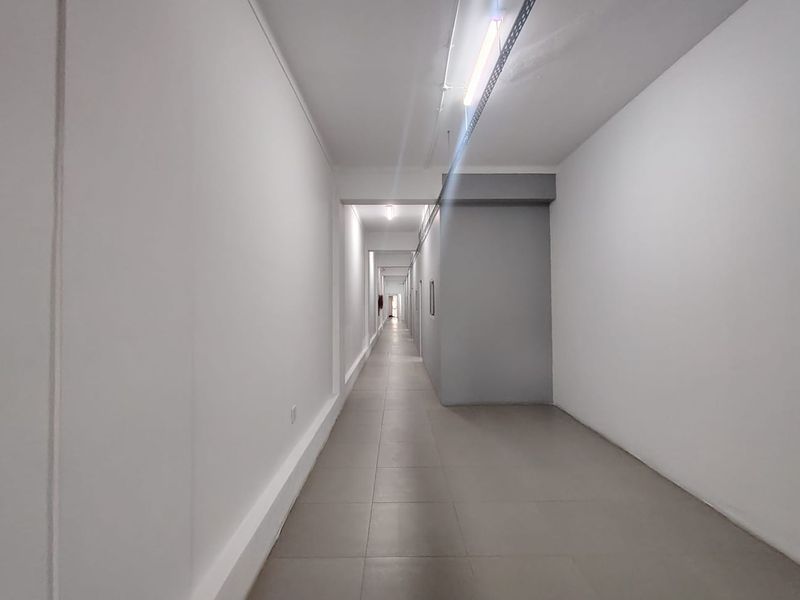A clean, minimalist office hallway featuring neutral tiled flooring, white walls, and exposed overhead lighting fixtures.