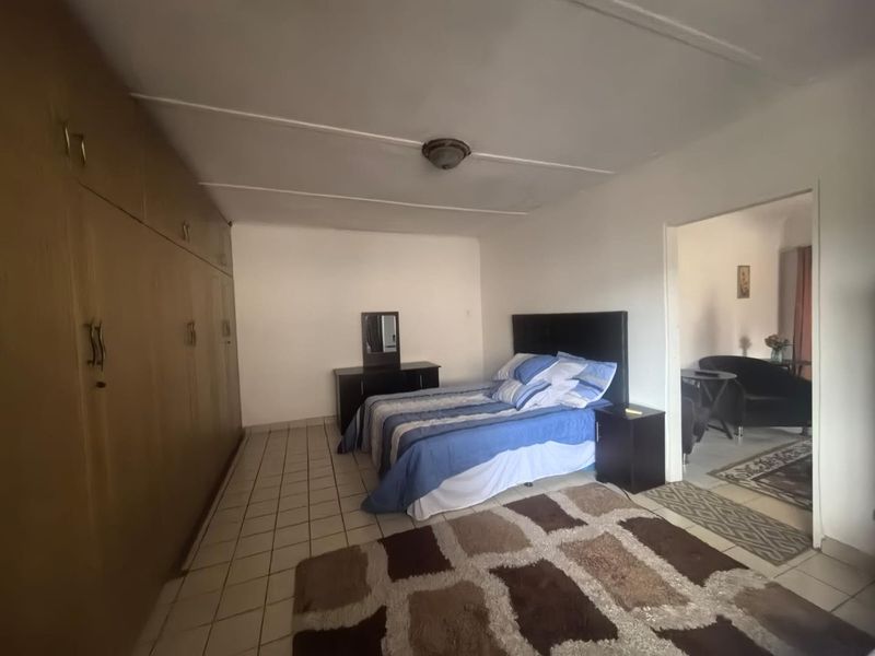 A bedroom featuring built-in wooden cupboards and tiled flooring, with an adjacent living area visible through the doorway.