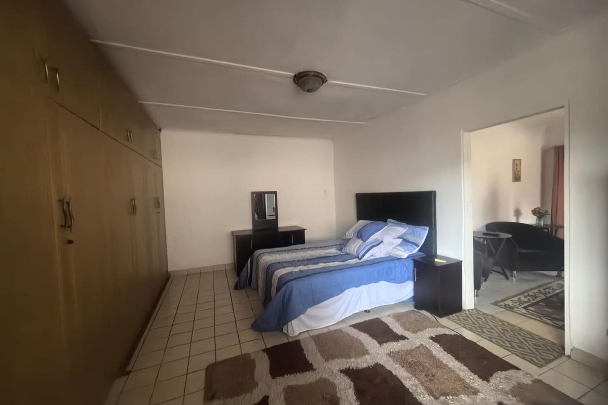 A bedroom featuring built-in wooden cupboards and tiled flooring, with an adjacent living area visible through the doorway.