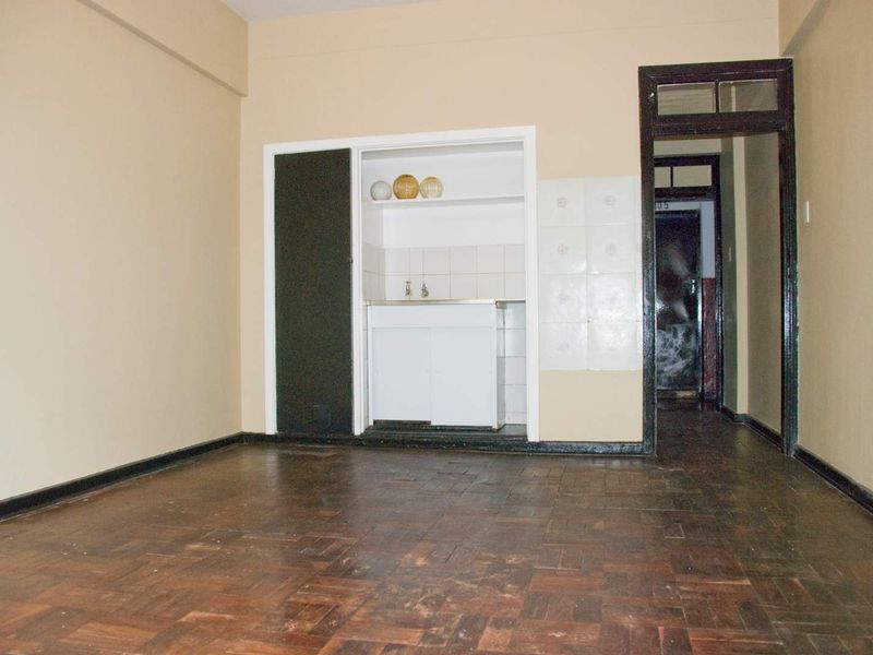 An empty living area featuring original parquet flooring and a compact built-in kitchenette with a sink and tiled splashback.