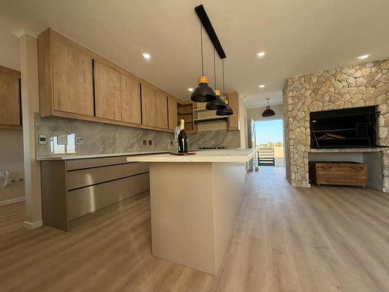 Modern open-plan kitchen featuring a central island, stone splashback, and a striking stone-clad fireplace with a built-in braai, leading to an outdoor area.