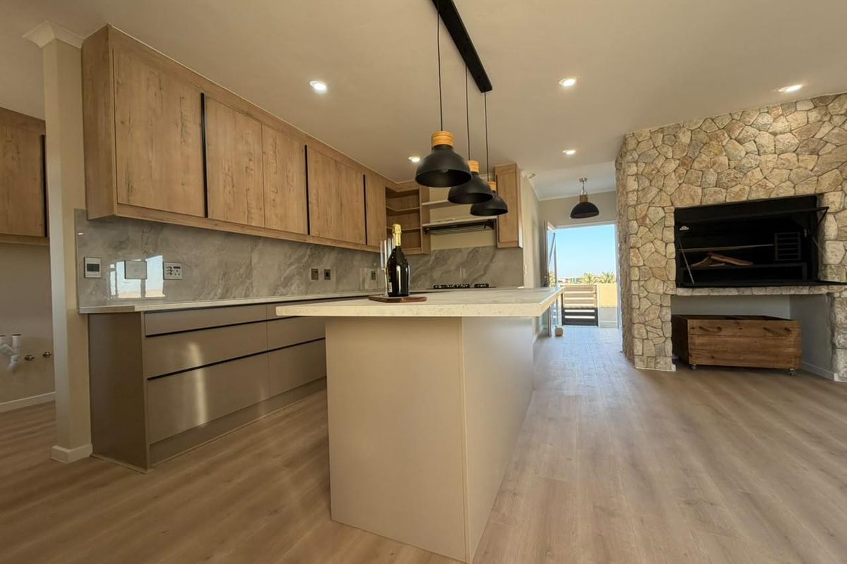 Modern open-plan kitchen featuring a central island, stone splashback, and a striking stone-clad fireplace with a built-in braai, leading to an outdoor area.