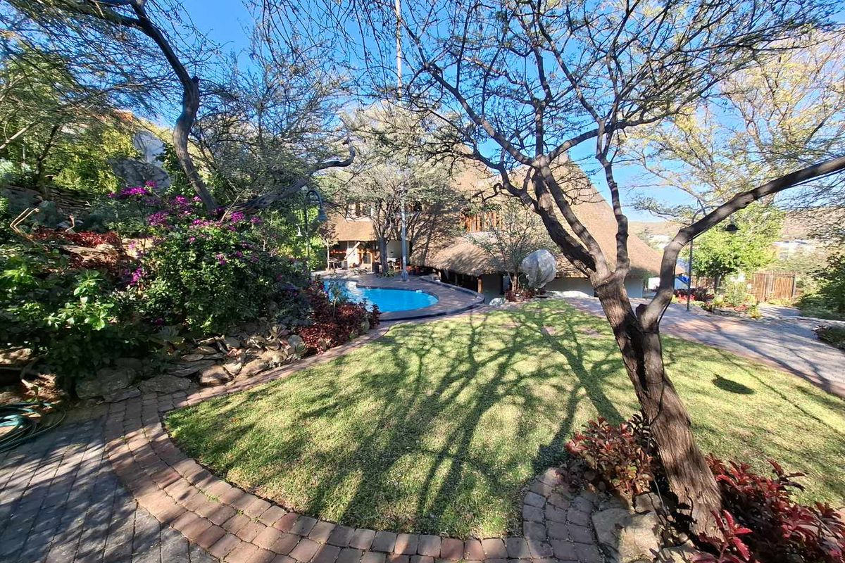 A landscaped rear garden featuring a swimming pool, paved pathways, and mature trees, set against the backdrop of a thatched-roof residence.