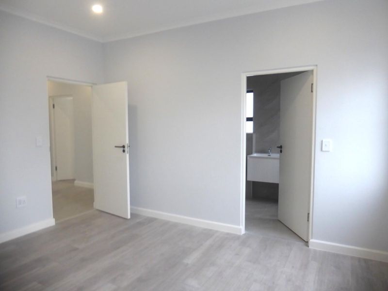 A bright, modern, and empty bedroom featuring light-toned laminate flooring, neutral walls, and direct access to an adjoining bathroom.
