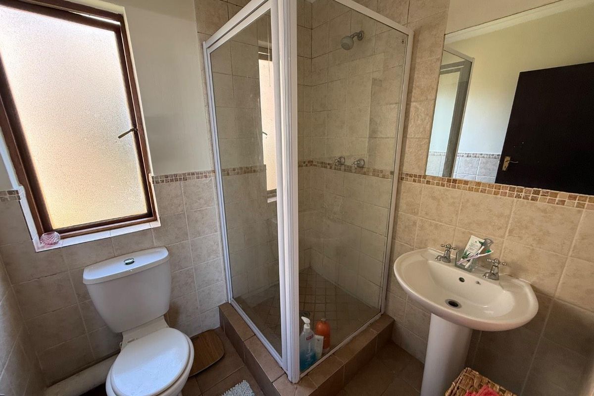 A functional bathroom featuring a tiled shower enclosure, pedestal basin, toilet, and neutral wall tiling with a frosted window for natural light.
