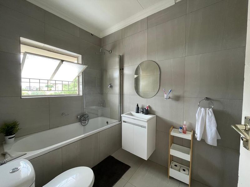 Modern bathroom featuring grey wall tiles, a bathtub with a glass shower screen, a wall-mounted vanity, and a toilet.