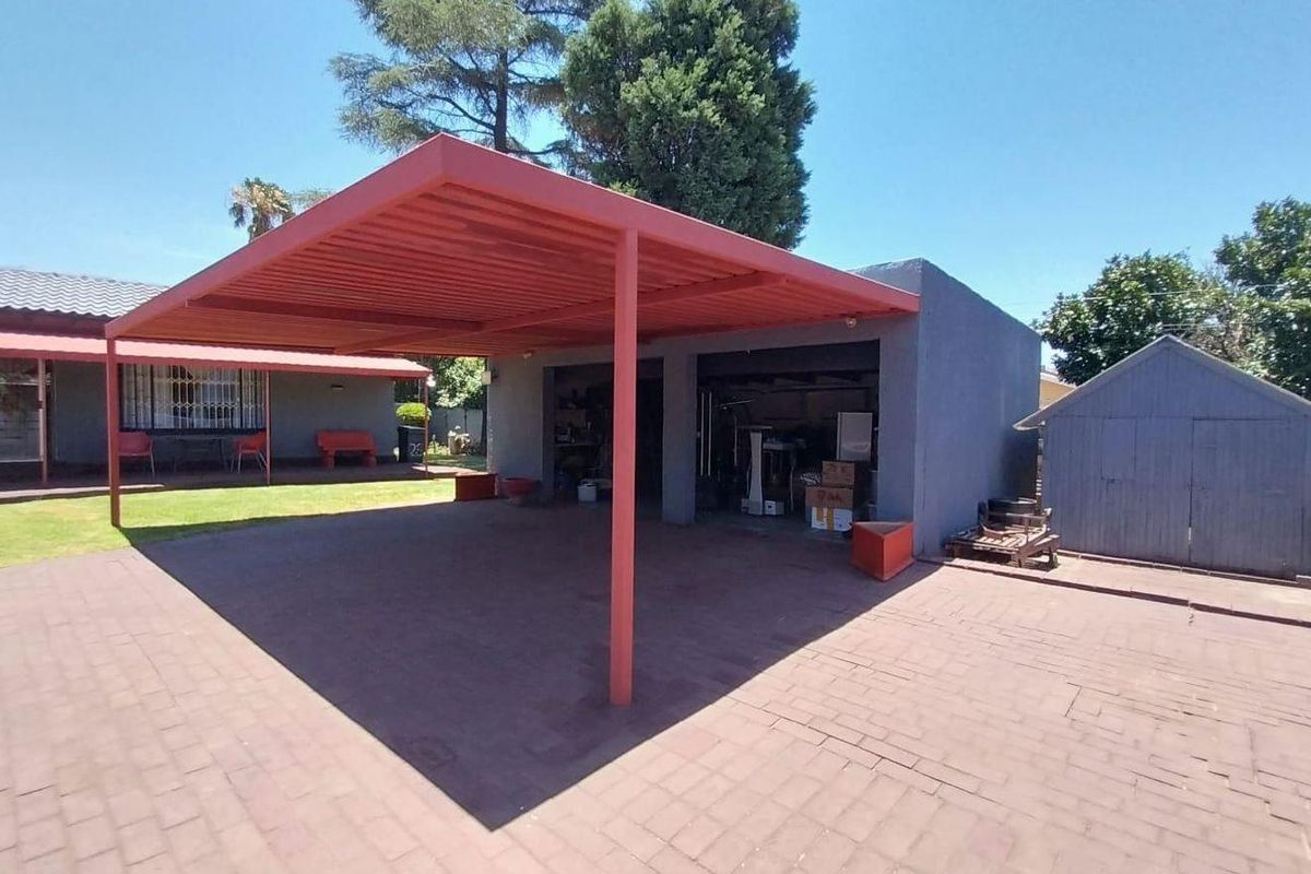 Rear exterior view featuring a paved driveway, a red metal carport, a double garage, and a detached garden shed set within a lawned area.