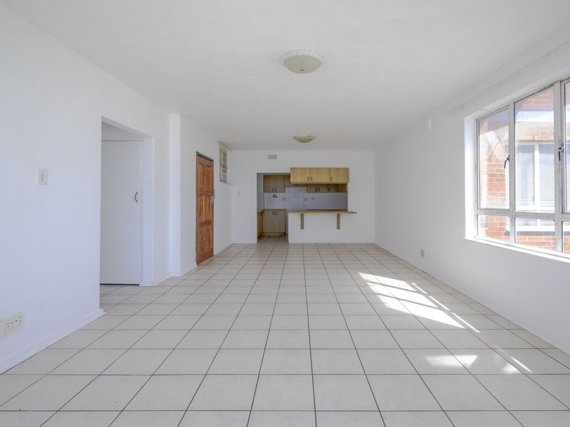 An empty open-plan living and kitchen area featuring tiled flooring, a breakfast bar, and natural light from a large window.